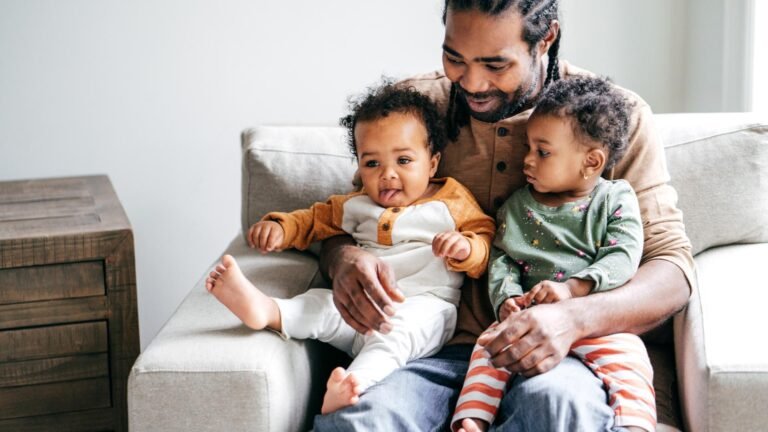 A smiling man sits on a beige couch holding two young children on his lap, sharing a playful moment. This warm scene could follow the peace of mind after receiving results from a court admissible DNA paternity test. A wooden table is beside them.