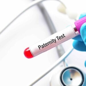 A gloved hand holds a blood sample tube labeled "Paternity Test," hinting at a DNA paternity testing kit. In the blurred background, a stethoscope suggests a medical or laboratory setting focused on paternity determination.