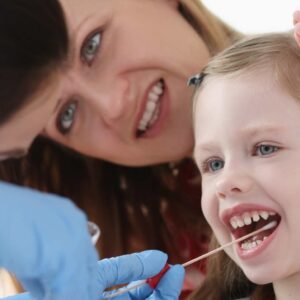 A smiling young girl with light skin and blue eyes has her mouth open for a cheek swab by a gloved hand. An adult woman, likely her mother, is closely watching, smiling encouragingly in the background. The setting appears to be a medical or clinical environment.