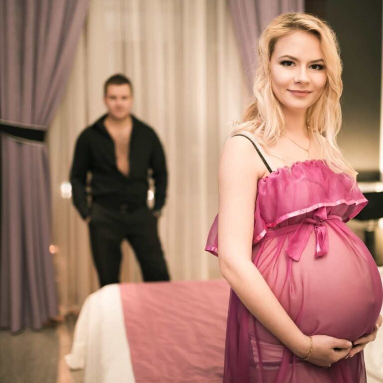 A pregnant woman in a sheer pink dress stands smiling near a bed, cradling her belly—embracing this new chapter as they consider a PRENATAL DNA PATERNITY TEST COURT ADMISSIBLE. Behind her, a man stands by the softly lit, purple-curtained room.