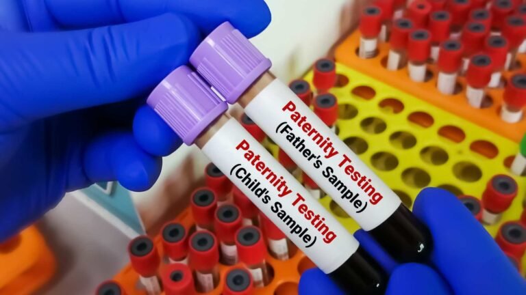 A close-up of two gloved hands holding blood sample tubes labeled "Paternity Testing (Father's Sample)" and "Paternity Testing (Child's Sample)," displaying the precision required for DNA court admissible paternity testing. Test tubes sit in yellow and orange racks behind.