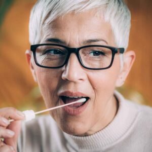 Close-up of an older woman with short gray hair and glasses, calmly swabbing the inside of her cheek, possibly for a legal relationship DNA test or other court admissible medical sample. She appears focused.
