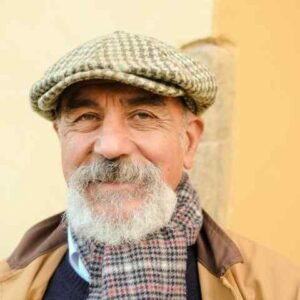 Smiling older man with a gray beard and mustache wearing a houndstooth flat cap and plaid scarf, perfect for a RELATIONSHIP DNA PATERNITY TESTING campaign. He has a tan jacket over a dark sweater; the softly focused background features yellow and beige tones.