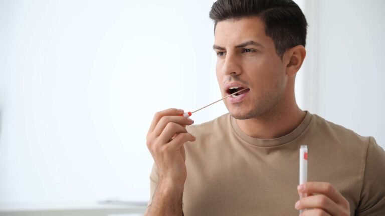 A man with short dark hair and a trimmed beard swabs his cheek with a cotton swab while holding a small plastic tube, likely for collecting a DNA sample for an AT HOME DNA PATERNITY TEST, in a bright, neutral indoor setting.