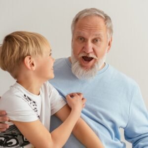 A smiling older man with white hair and a beard, wearing a light blue sweater, embraces a laughing young boy. Their joyful moment reflects the special bond explored through RELATIONSHIP DNA PATERNITY TEST, set against a plain, light-colored background.